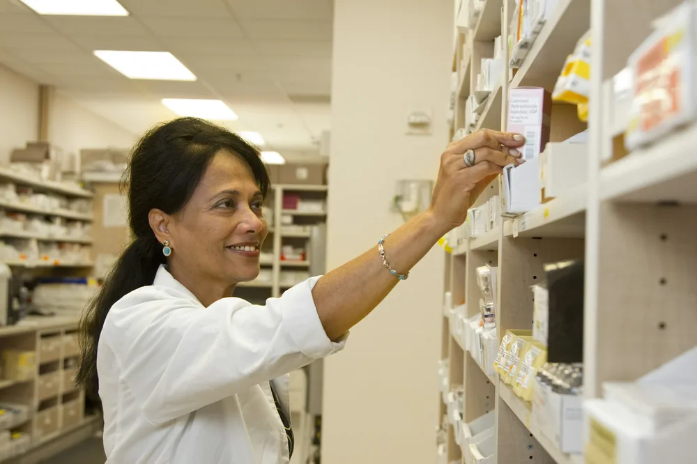 A pharmacist in a white coat selecting a medication carton from pharmacy shelving